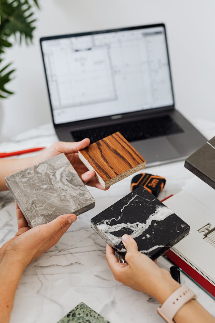 Hands assessing diverse tile samples on marble table with laptop plans.